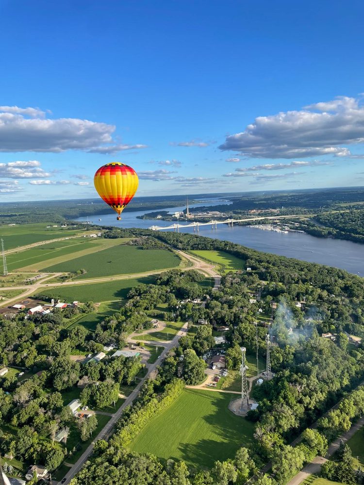 A red and yellow hot air balloon over green farmland with the St. Croix Crossing bridge and river in the distance